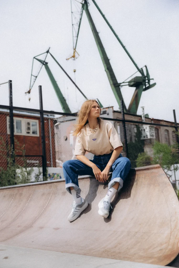 Kobieta w koszulce Miejska Fala siedzi na rampie skateparku, w tle dźwigi stoczniowe